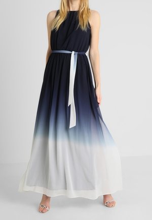 Kobieta w bezrękawną, sięgającą podłogi suknię ombre, przechodzącą z granatowego do białego, przewiązaną w talii granatowo-białym paskiem.