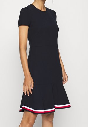Kobieta w krótkiej sukience w kolorze granatowym z rozkloszowanym dołem i detalem w paski w kolorze czerwonym, białym i granatowym na krawędzi.