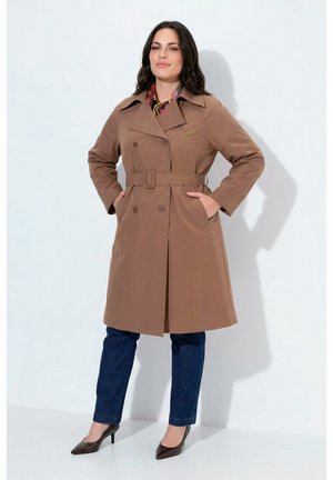 Donna in piedi con le mani nelle tasche, indossa un trench coat marrone con cintura, jeans blu scuro e scarpe con tacchi alti scuri su sfondo bianco.