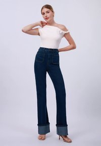 Jonge vrouw poseert in een off-shoulder witte top, high-waisted donkerblauwe jeans met grote omslagen, en open teen hakken tegen een effen achtergrond.