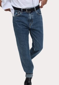 Vaqueros de mezclilla azul oscuro con un corte relajado, puños enrollados, cinco bolsillos y un cinturón negro con una hebilla rectangular de plata.