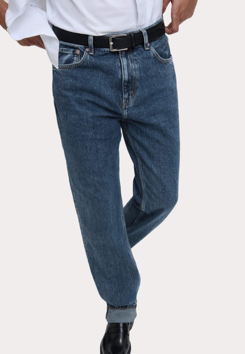 Vaqueros de mezclilla azul oscuro con un corte relajado, puños enrollados, cinco bolsillos y un cinturón negro con una hebilla rectangular de plata.