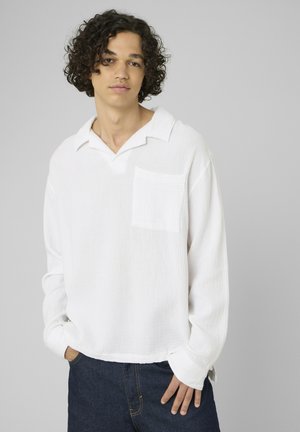 Jeune personne aux cheveux bouclés portant une chemise blanche ample à manches longues et un jean foncé, debout avec une main dans la poche devant un fond gris.