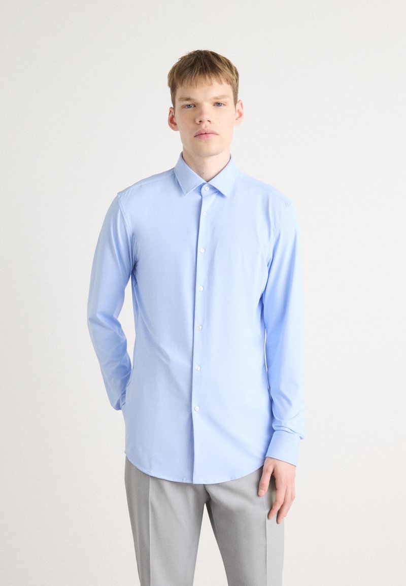 Camisa azul clara de manga larga con cuello abotonado, textura suave y botones blancos, combinada con pantalones gris claro.
