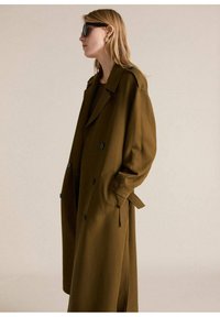 Trench coat verde oliva con un diseño de doble botonadura, bolsillos laterales y un cinturón para atar. Presenta una textura suave y un corte oversize.