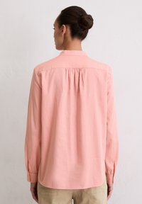 Light pink long-sleeved shirt with a back yoke and gathered detail, featuring a collar and buttoned cuffs. Soft, smooth fabric texture.