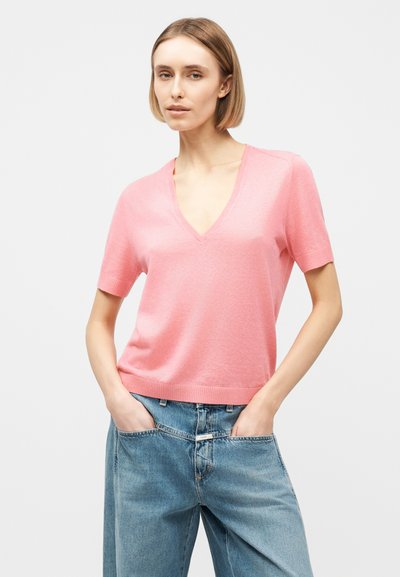 Femme aux cheveux blonds courts portant un pull rose à manches courtes en V et un jean bleu ample, les mains dans les poches, sur un fond uni.