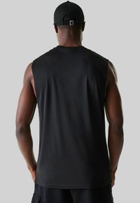 Mann in schwarzem ärmellosem Shirt und schwarzer Mütze, von hinten vor einem schlichten grauen Hintergrund gezeigt.
