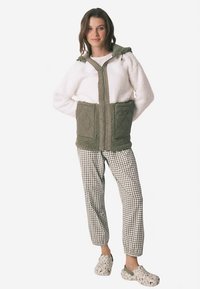 Valkoinen, pörröinen vetoketjullinen takki, jossa oliivinvihreitä tilkkuja ja huppu, yhdistetty mustavalkoisiin ruutukuviollisiin housuihin ja kuvioituihin puukenkäisiin.