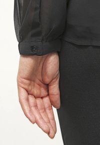 Main gauche détendue sur le côté, portant un chemisier noir transparent à manches longues avec des poignets boutonnés et un pantalon noir.
