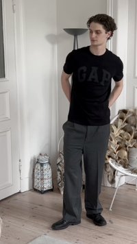Camiseta de algodón negra con el logo de "GAP", pantalones grises de corte holgado, zapatos negros, de pie en una habitación luminosa con decoración minimalista.