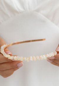 Opaska do włosów w złotym odcieniu z gładkimi, owalnymi kamieniami przypominającymi opale w jasnych odcieniach, ułożonymi z przodu dla efektu wizualnego.