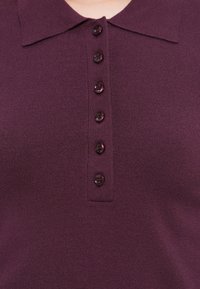 Soeproheline polo-särk sügavas lillakas värvitoonis, millel on klassikaline krae ja kuus tumedat nuppu sanga ääres. Sile tekstuur ja keha lähedane siluett.