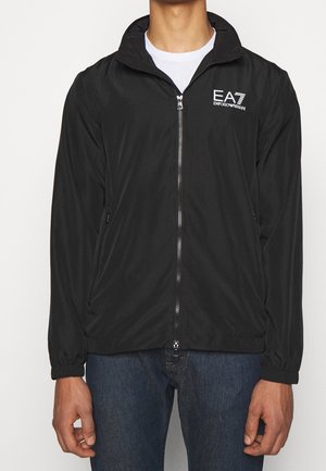 Mężczyzna ubrany w czarną kurtkę zapinaną na zamek z logo EA7 Emporio Armani na piersi, w połączeniu z niebieskimi jeansami i białą koszulą pod spodem.