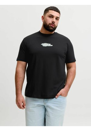 Homme barbu portant un t-shirt noir à motif graphique et un jean bleu clair, debout avec une main dans la poche devant un fond uni.