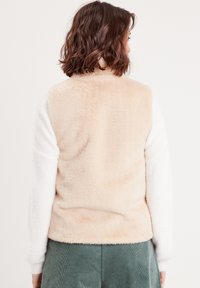 Gilet en fausse fourrure beige au toucher doux, avec des manches longues blanches et un ourlet arrondi. La vue arrière met en avant un design simple et épuré.
