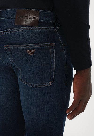 Σκούρο μπλε τζιν παντελόνι με δερμάτινη ετικέτα στη μέση που φέρει �ανάγλυφο όνομα μάρκας. Τσέπη με ραφή λογότυπου και αντίθετο νήμα.