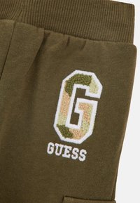 Pantalons de surpoids vert kaki avec ceinture côtelée, ornés d'un logo "G" en broderie dans des couleurs camouflage et du texte blanc épelant "GUESS."