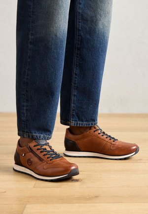 Baskets en cuir marron avec des lacets et des semelles noires portées avec un jean bleu sur un sol en bois clair.