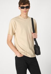 T-shirt en coton beige avec une coupe décontractée, associé à un pantalon noir et un petit sac à épaule noir, porté avec des lunettes de soleil noires rectangulaires.