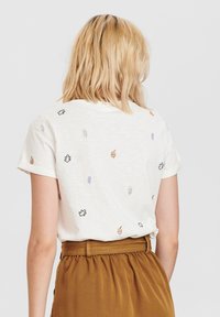 Kobieta z blond włosami do ramion, ubrana w białą koszulę z krótkim rękawem z małymi wzorami liści oraz brązową spódnicę o wysokim stanie, odwrócona plecami.