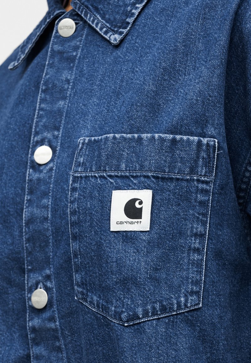 Denim shirt in dark blue with a button front, pointed collar, chest pocket, and a Carhartt logo tag. Features silver-tone buttons.
