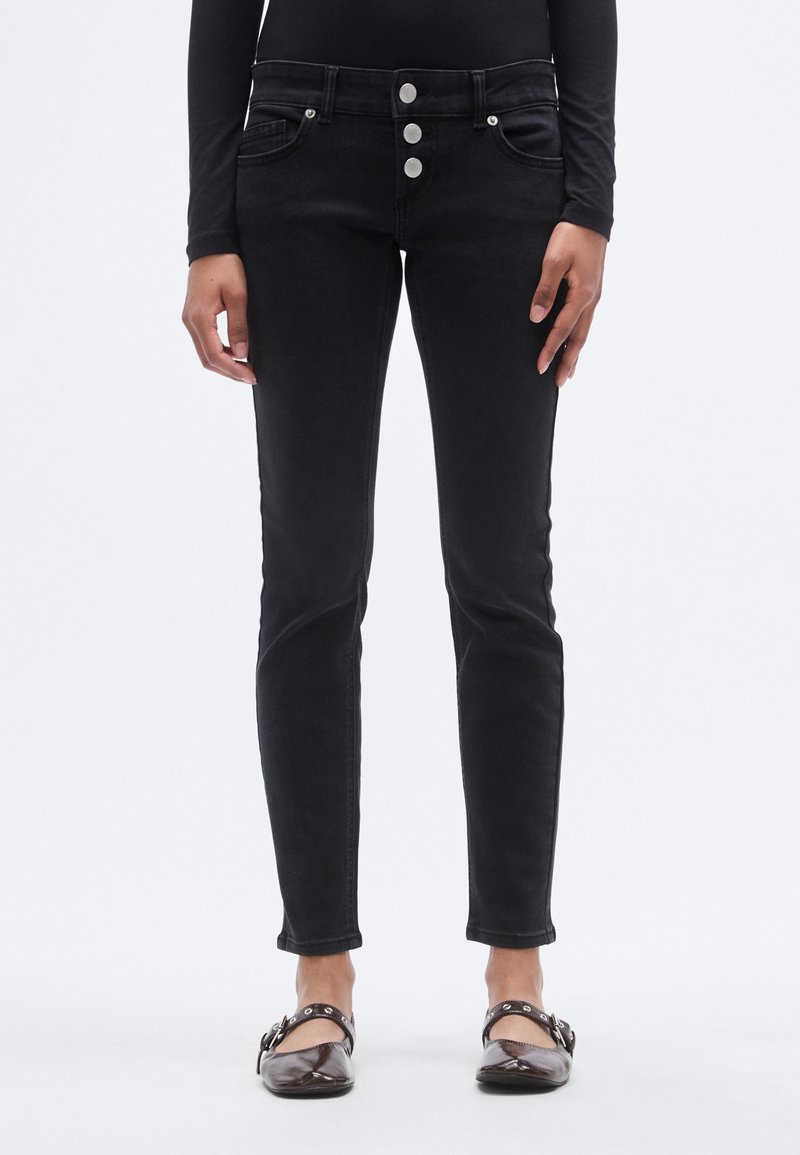 Μαύρο τζιν skinny με ψηλή μέση, τρία μεταλλικά κουμπιά μπροστά και αντίθετες ραφές. Το μοντέλο φοράει σκουρόχρωμα φλατ παπούτσια.