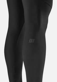 Leggings neri realizzati in materiale texturizzato, con un design aderente e un piccolo logo vicino al ginocchio. La superficie è liscia con una leggera lucentezza.