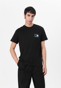 T-shirt noir en coton à manches courtes, avec un petit graphique multicolore sur la poitrine gauche et un col rond.