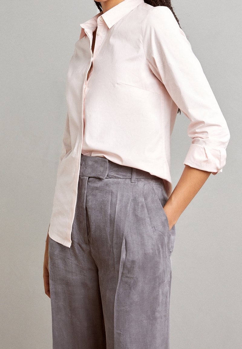 Femme portant une chemise rose clair à boutons avec les manches retroussées et un pantalon gris large taille haute, les mains dans les poches.