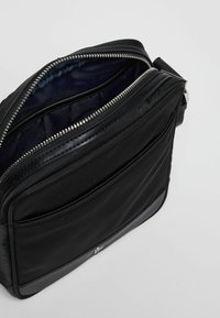 Borsa a tracolla nera in tessuto con scomparto principale con zip, texture liscia e un accento di hardware lucido. L'interno è di colore blu navale.