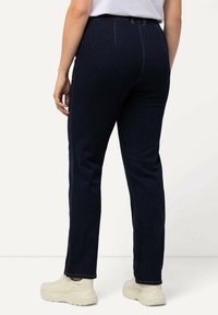 Jeans de mezclilla azul oscuro con un diseño de pierna recta, estilo de cinco bolsillos y costuras naranjas contrastantes, combinados con zapatillas de color claro.