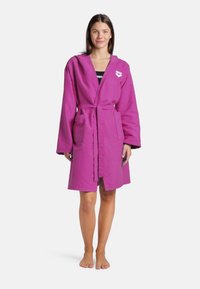 Accappatoio viola in cotone con cappuccio, maniche lunghe, cintura in vita e tasche frontali. Presenta un logo sulla parte sinistra del petto.