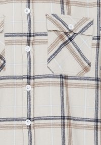 Beige geblokt shirt van flanel; heeft bruine, marineblauwe en witte lijnen; bevat twee borstzakken en witte knopen.