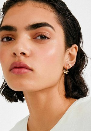 Kuldsetest lillelistest kõrvarõngastest koosnev väike lilledega disain ja rõngas. Mudelil on tume, laineline juuksed ja loomulik meigistiil.