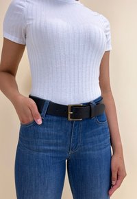 Svart läderbälte med en guldfärgad metallspänne, buren över bl�å denimjeans och en vit, ribbade kortärmad topp. Material och texturer synliga.