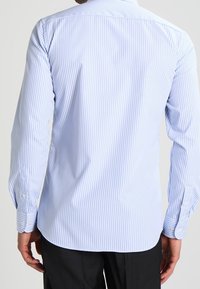 Een man die een lichtblauw en wit verticaal gestreept overhemde met lange mouwen draagt, bekeken vanaf de achterkant, gecombineerd met zwarte broeken.