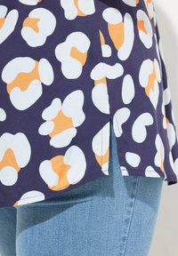 Top blu navy con un motivo astratto bianco e arancione, caratterizzato da una fessura laterale e un orlo cucito, abbinato a jeans azzurri chiari.