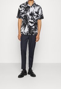 Chemise à manches courtes avec col, imprimé de palmiers noirs sur fond blanc, associée à un pantalon ajusté foncé et des chaussures de ville noires.