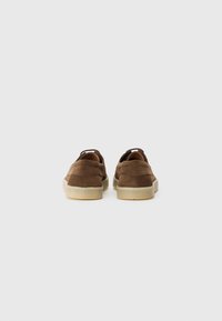 Brune semskede slip-on sko med en glat, lysfarget gummisåle og minimale sømdetaljer, sett fra baksiden.