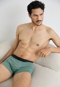 Tumši zaļi rakstaini bokseršorti ar melnu elastīgu jostu, ar ģeometrisku dizainu, ko valkā vīriešu modelis, sēžot uz teksturētiem krēmkrāsas dīvāna.