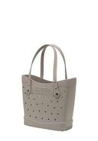 Sac fourre-tout en silicone beige avec un design perforé, poignées doubles et une base texturée. Marque affichée de manière proéminente à l'avant.