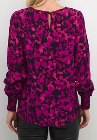 Blomstret bluse i sort med livlige pink og lilla mønstre. Har lange puffærmer, smockede manchetter og en nøglehulåbning i ryggen.
