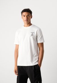 Witte katoenen t-shirt met een ronde halslijn, korte mouwen en een grafische print in het zwart op de linkerborst. Gecombineerd met zwarte broek.