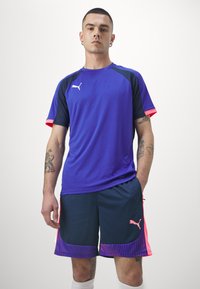 Puma Sportoutfit mit einem lila T-Shirt mit kurzen Ärmeln und schwarzen Akzenten sowie einer navyblauen Shorts mit rosa Highlights und einem gemusterten Saum.