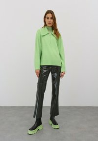 Mintgrüner Reißverschluss-Pullover mit einem großen Kragen, kombiniert mit schwarzen glänzenden Lederhosen und schwarzen Stiefeln mit dicker Sohle und limettengrünen Sohlen.