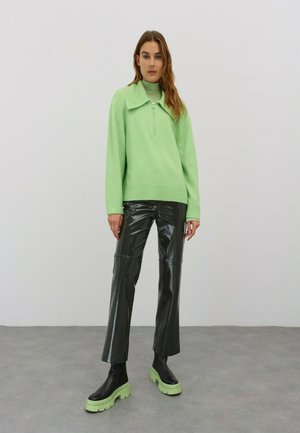 Mintgrüner Reißverschluss-Pullover mit einem großen Kragen, kombiniert mit schwarzen glänzenden Lederhosen und schwarzen Stiefeln mit dicker Sohle und limettengrünen Sohlen.