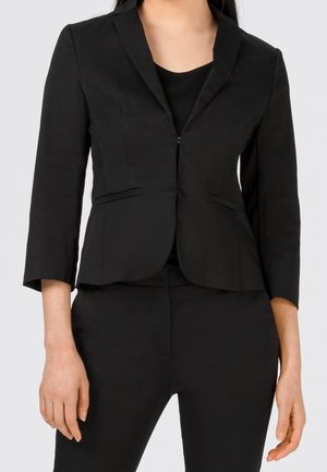 Kvinde iført en tætsiddende sort blazer med 3/4-ærmer over en sort top og sorte bukser, stående mod en ensartet lys baggrund.