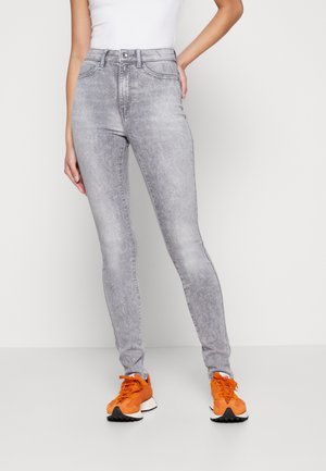 Femme portant un jean skinny taille haute gris clair et des baskets orange vif, debout avec une main sur la hanche sur fond blanc.