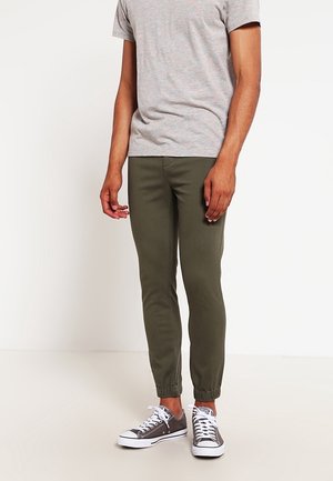 Mann trägt olivgrüne Jogginghosen, graues T-Shirt und graue Low-Top-Sneaker, steht vor einem weißen Hintergrund.